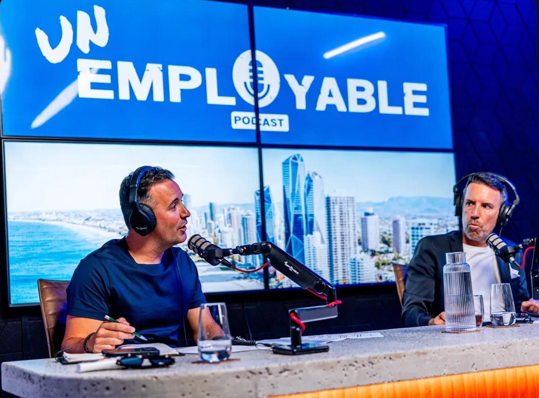 Two hosts wearing headphones at a studio desk, recording an Unemployable podcast session in front of a screen showing the Unemployable Podcast logo